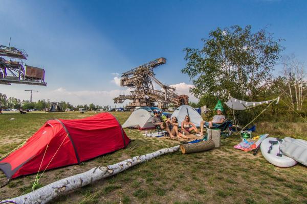 vier Personen sitzen auf einer Decke vor Campingzelten, im Hintergrund Ferropolis